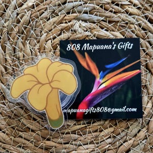 May include: A yellow flower sticker with a green stem. The sticker is on a woven background with a business card that says "808 Mapuana's Gifts" and "mapuanagifts808@gmail.com".