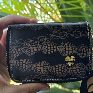 May include: Black wallet with a gold zipper and white stitching. The wallet features a repeating pattern of gold seashells. A gold logo with text is visible on the front.