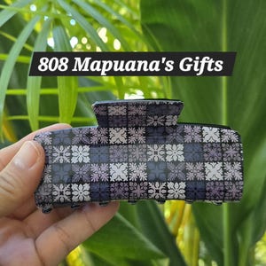 May include: A patterned hair claw clip in shades of gray, purple, and white. The clip features a geometric floral design and the text "808 Mapuana's Gifts" in a white font on a black background.
