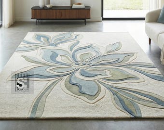 May include: A rectangular area rug featuring a large, central floral motif in shades of blue, sage green, and cream. The design includes layered, sculpted petals and leaves with a textured finish. A small black logo with the text SAJDA HOME is visible.