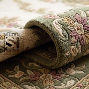 Puede incluir: Primer plano de una alfombra enrollada con un dise&ntilde;o floral. La alfombra presenta un fondo crema con patrones florales verdes y rosas. Los bordes son de color verde oscuro con un borde decorativo. La marca "SAJDA HOME" es visible.