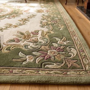 Puede incluir: Una alfombra ornamental con un centro color crema y un borde verde. La alfombra presenta dise&ntilde;os florales y de volutas en tonos verdes, rosas y beige. La alfombra est&aacute; colocada sobre un suelo de madera, con parte de una silla visible.