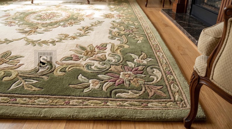Puede incluir: Una alfombra ornamental con un centro color crema y un borde verde. La alfombra presenta dise&ntilde;os florales y de volutas en tonos verdes, rosas y beige. La alfombra est&aacute; colocada sobre un suelo de madera, con parte de una silla visible.