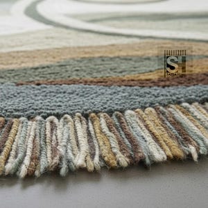 Puede incluir: Primer plano de una alfombra multicolor con una superficie texturizada y un borde con flecos. La alfombra presenta rayas horizontales en tonos marrones, verdes y grises, con un dise&ntilde;o de remolino crema. La alfombra est&aacute; hecha de un material suave y tejido.
