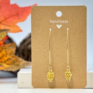 May include: Gold-colored dangle earrings with a pinecone design. The earrings hang from a long, thin gold bar. The earrings are displayed on a brown card with the word "Handmade" and a heart symbol.