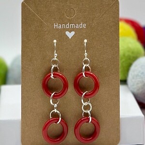 May include: Red hoop earrings with silver-tone metal links, displayed on a brown card with the word "Handmade" and a heart symbol. The earrings feature three connected red hoops.
