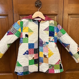May include: A handmade, quilted jacket with a patchwork design. The jacket features a variety of colorful fabric squares, including red, blue, green, and yellow. It has a black zipper and a red patterned hood. The jacket is hanging on a wooden hanger.