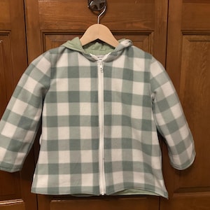 May include: A hooded jacket with a green and white plaid pattern. The jacket has a full-length white zipper and a light green lining. The jacket is hanging on a wooden hanger.