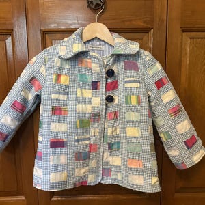 May include: A light blue quilted jacket with a gingham pattern and colorful rectangular patches. The jacket has a rounded collar, long sleeves, and two dark blue buttons. The jacket is hanging on a wooden hanger.