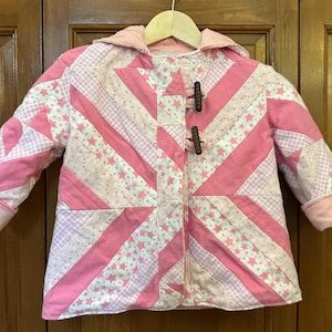 May include: A pink and white quilted jacket with a hood and toggle closures. The jacket features a patchwork design with pink stripes, stars, and gingham patterns. The cuffs and hood lining are solid pink.