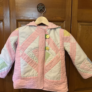 May include: A quilted jacket in pastel pink, white, and green patchwork. The jacket features a hood, button closure, and a variety of patterned fabrics. The jacket is hanging on a wooden hanger.