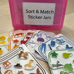 May include: A pink plastic box labelled "Sort & Match Sticker Jars" contains cards with sticker images. The cards display stickers of a tiger, a star, a heart, a butterfly, and a whale. The stickers are designed for sorting and matching activities.
