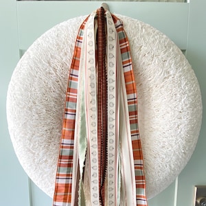 May include: A circular wreath made of white textured material adorned with a collection of hanging ribbons. The ribbons feature various patterns including orange and green plaid, solid brown, and a cream ribbon with a repeating pumpkin print.