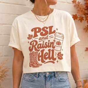 May include: A cream-colored short-sleeve t-shirt featuring a rust-colored graphic with the text PSL and Raisin Hell. The design includes a coffee cup with a pumpkin icon, a pair of cowboy boots, and maple leaves, all printed in a retro font style.