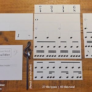 May include: A set of 40 white rhythm tiles with black musical notes and symbols. The tiles are arranged in rows on a wooden surface. The tiles are labelled with numbers and include a booklet titled "Music Unpacked Rhythm Builder".