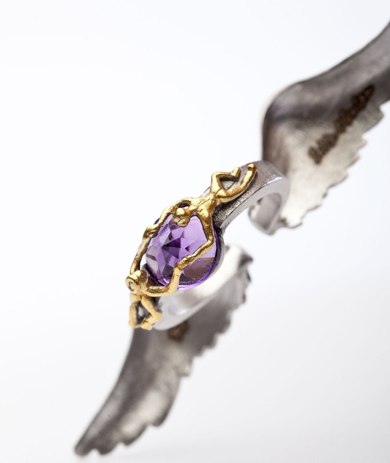 May include: A silver ring with a gold filigree design and a large, oval-cut purple gemstone. The ring is held by a silver wing-shaped element, creating a unique and artistic presentation. The ring's design is intricate and eye-catching.