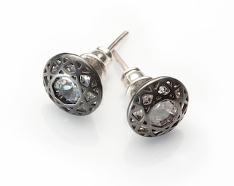 May include: Two silver-toned stud earrings with a dark gray, openwork design around a central, clear gemstone. The earrings have a post backing and are set against a white background.