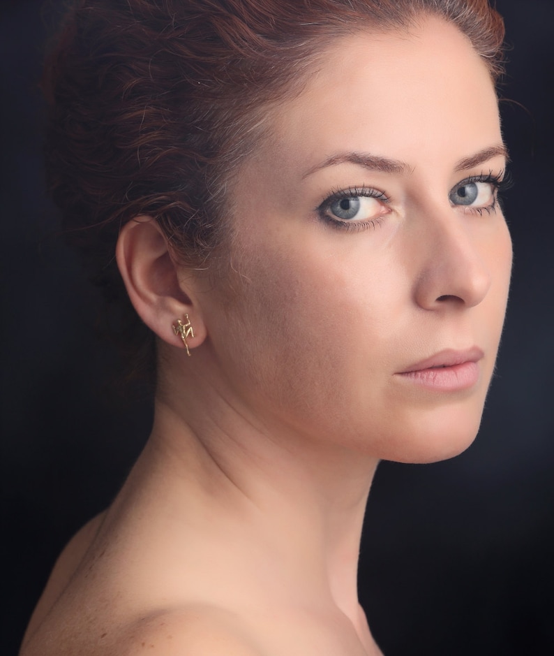 May include: A woman with red hair and blue eyes is wearing a small gold earring in the shape of a person climbing a rope.