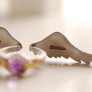 May include: A silver ring with a purple gemstone and two silver wing-shaped earrings. The earrings have the text "Selda Okutan" engraved on them. The ring has a gold-colored band.