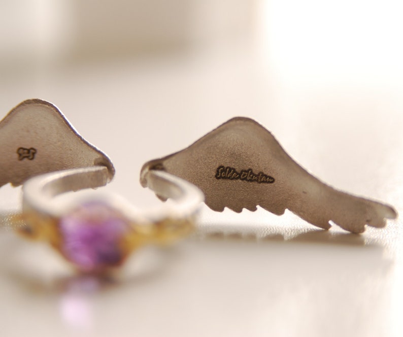 May include: A silver ring with a purple gemstone and two silver wing-shaped earrings. The earrings have the text "Selda Okutan" engraved on them. The ring has a gold-colored band.