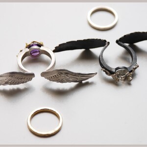 May include: Several rings with unique designs are displayed. One ring features a purple gemstone and gold accents. Two rings have wing-shaped elements. Two plain bands are also present. The rings are silver and black.