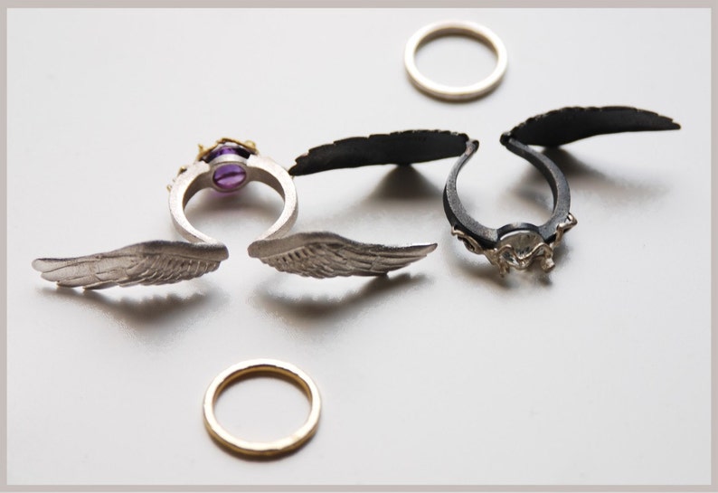 May include: Several rings with unique designs are displayed. One ring features a purple gemstone and gold accents. Two rings have wing-shaped elements. Two plain bands are also present. The rings are silver and black.
