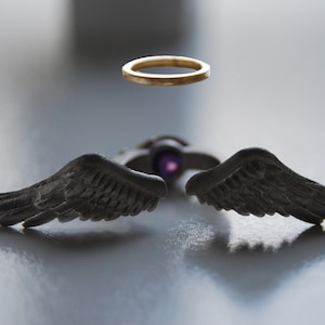 May include: A unique jewelry set featuring a gold halo ring and a pendant with dark gray angel wings. The pendant has a purple center stone. The wings are detailed with feather textures, creating a striking contrast.