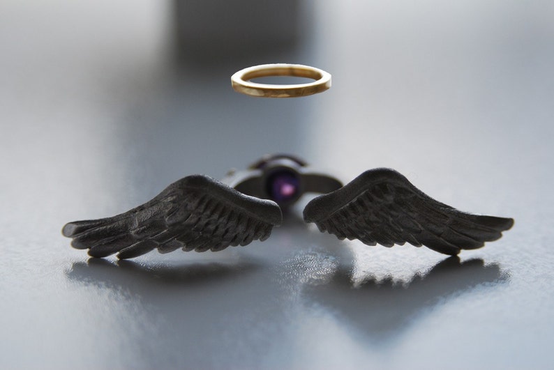 May include: A unique jewelry set featuring a gold halo ring and a pendant with dark gray angel wings. The pendant has a purple center stone. The wings are detailed with feather textures, creating a striking contrast.