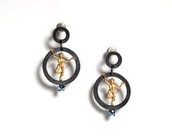 May include: A pair of unique earrings featuring a gold-colored figure within a dark gray circular frame. Each earring has a small blue gemstone and a smaller circular accent above. The earrings are set against a white background.
