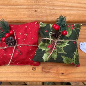 May include: Two small, fabric-wrapped gifts tied with twine. One is red with gold stars and the other is green with a holly pattern. Both are decorated with red berries and a small pine cone. The green gift has a tag that says "cinnamon roll".