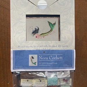 May include: A close-up of a Mermaid Verde embellishment pack for cross-stitch. The pack includes a variety of glass seed beads in different colors, including green, red, orange, and blue. The pack is labeled with the name "Nora Corbett" and the product number "NC192E".