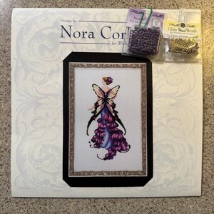 May include: A cross-stitch kit featuring a framed design of a fairy with purple and black accents. The kit includes beads and is designed by Nora Corbett.