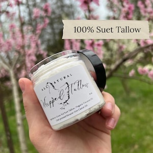 May include: A clear jar of whipped tallow with a black lid, held in a hand. The label reads "Whipped Tallow" and "All Natural". The jar is 4 oz. The background is blurred with pink flowers and the text "100% Suet Tallow".