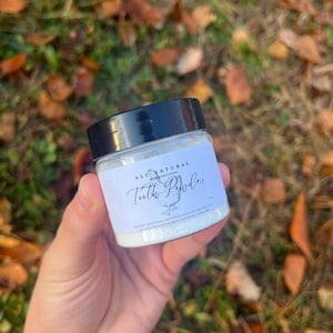 May include: A small white jar with a black lid containing a white powder. The jar has a label that says "All Natural Remineralizing Tooth Powder".