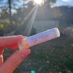 May include: A white tube of lip balm with the text "Honey Tallow Lip Balm" and a floral design. The tube is being held in a hand against a blurred background of trees and a bright sun.