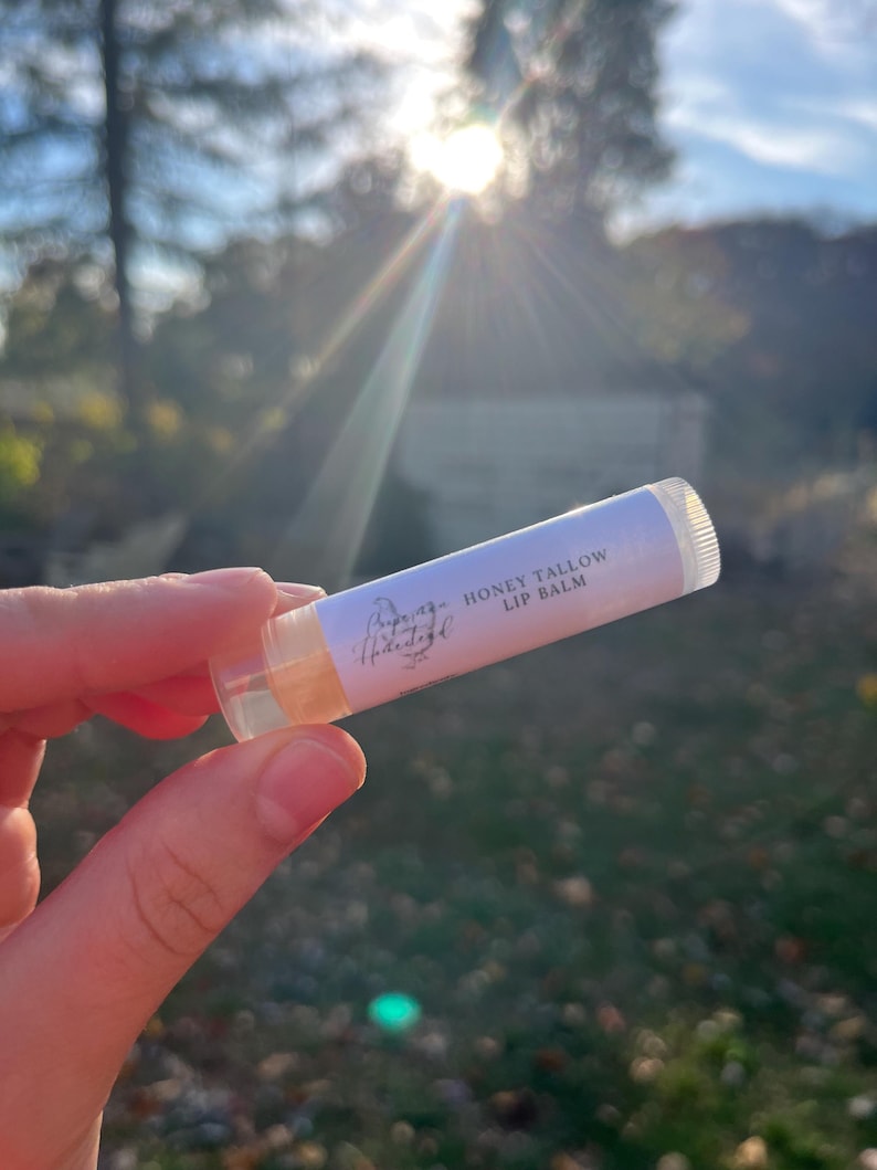 May include: A white tube of lip balm with the text "Honey Tallow Lip Balm" and a floral design. The tube is being held in a hand against a blurred background of trees and a bright sun.