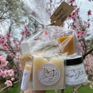 May include: A clear plastic gift bag filled with bath and body products. The bag has a brown tag that says "happy mother's day". The products include a bar of soap, a jar of whipped body butter, a small bottle of lotion, a loofah, and a lip balm. The products are all natural and handmade.