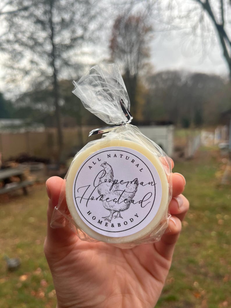 May include: A round, white soap bar with a white and black label that says "All Natural Cooperman Homestead Home & Body" with a chicken illustration.