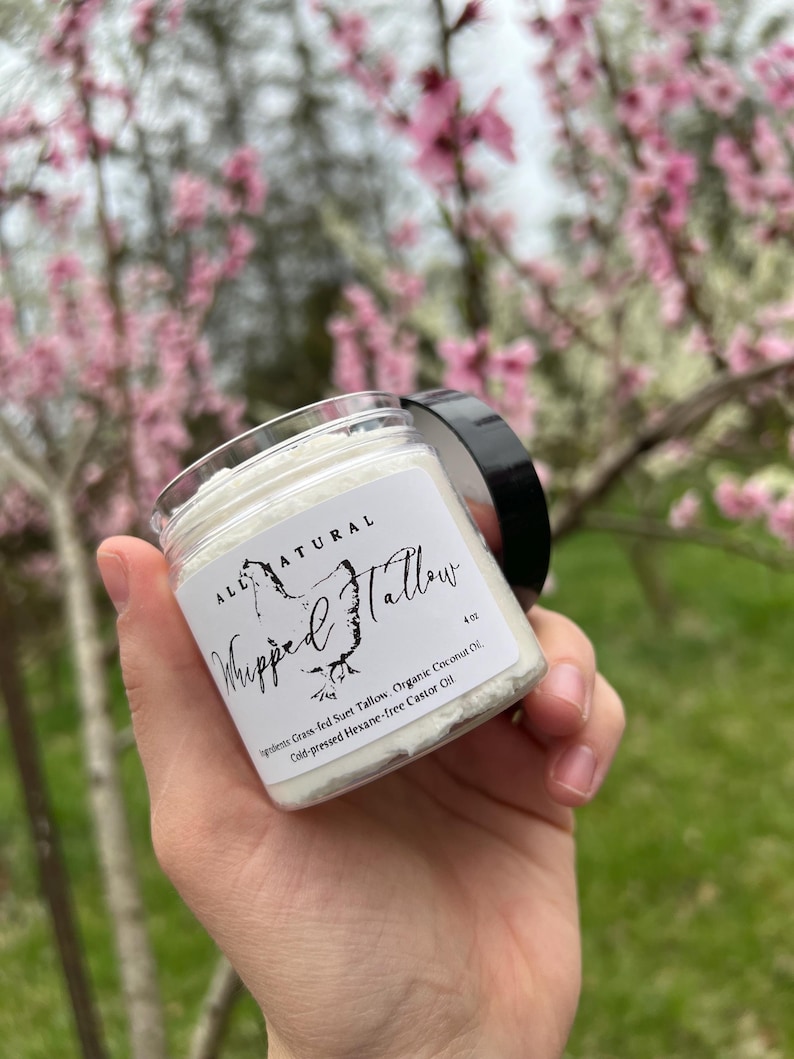 May include: A white jar with a black lid labeled "ALL NATURAL Whipped Tallow 4 oz" with the ingredients listed: "Grass-fed Suet Tallow, Organic Coconut Oil, Cold-pressed Hexane-free Castor Oil".