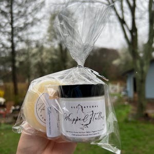 May include: A clear cellophane bag contains a jar of "Whipped Tallow", a lip balm, and a round bar of soap. The jar has a black lid and a white label with black text. The soap is yellow and the lip balm is in a small tube.