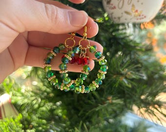 May include: Green beaded hoop earrings with gold accents and small red bead clusters. The earrings are held in front of a Christmas tree, with a white ornament in the background. A festive accessory.