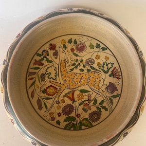 Può includere: Una ciotola rotonda in ceramica con interno color crema caratterizzata da un'illustrazione centrale di un cervo maculato circondato da fiori e fogliame colorati. L'ampio bordo smerlato è decorato con una fascia scura e piccoli motivi floreali.