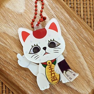 May include: A white Maneki-neko cat keychain with red accents on its ears and head. The cat holds a gold coin and a scroll. The keychain has a red ball chain and is on a wooden tray.