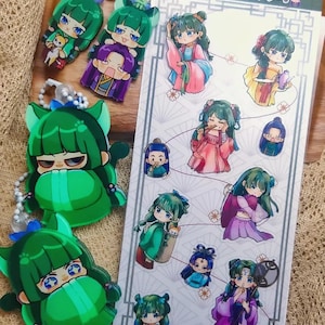 Op de afbeelding: Een verzameling sleutelhangers en stickers met anime-personages. De sleutelhangers zijn groen met blauwe accenten en goudkleurige hardware. De stickers tonen verschillende personages in kleurrijke traditionele kleding. De tekst op het stickervel is in het Japans.