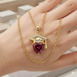 Può includere: Collana color oro con un ciondolo a forma di cappello di laurea. Il ciondolo ha una pietra preziosa rosso scuro a forma di cuore e il nome "Sofia" in bianco. La collana è su una mano.
