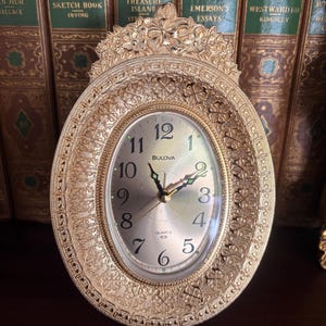May include: A gold-toned Bulova quartz clock with an oval face and ornate filigree detailing. The clock face features black numerals and hands, with the brand name "BULOVA" at the bottom. The clock is set against a backdrop of antique books.