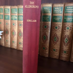 May include: A maroon book titled "The Allinghams" by Sinclair, with the Macmillan publisher's mark at the bottom. The book is standing upright on a shelf with other antique books in the background.