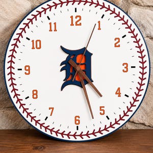 May include: A round wall clock with a white face featuring a red baseball stitch pattern along the perimeter. The clock has orange numbers from 1 to 12, a dark blue and orange Detroit Tigers logo in the center, and three dark brown wooden hands.