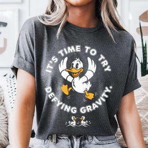 Puede incluir: Camiseta gris oscuro con un dibujo de un pato de dibujos animados y el texto "IT'S TIME TO TRY DEFYING GRAVITY." El pato tiene pico y patas amarillas. La camiseta es de estilo casual.