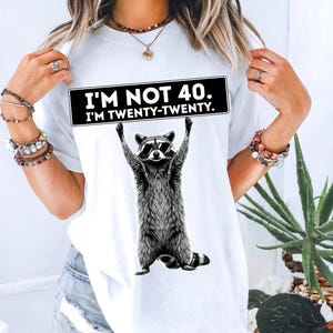 Puede incluir: Camiseta blanca con una ilustración en blanco y negro de un mapache con gafas de sol que sostiene un letrero que dice "I'M NOT 40. I'M TWENTY-TWENTY."
